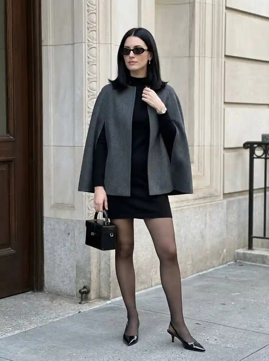 Woman in a charcoal cape layered over a black mini dress with sheer tights, black slingback heels, and a structured handbag.