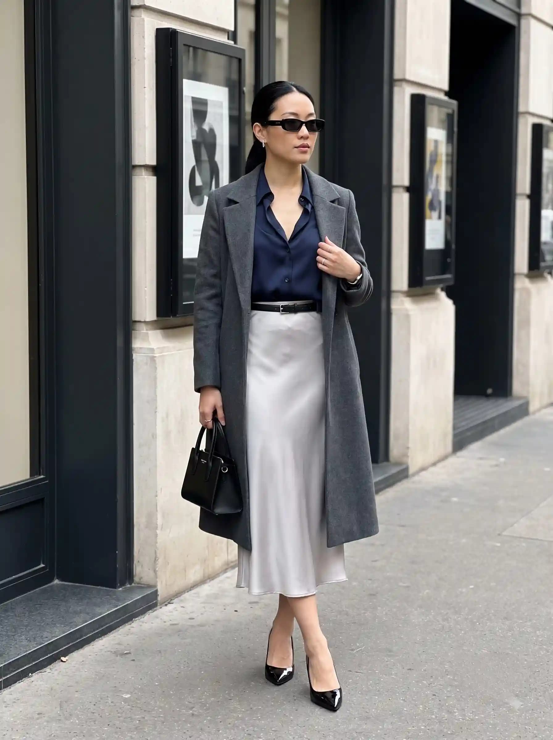 Woman wearing a charcoal long coat with a navy blouse, silver satin midi skirt, black belt, structured handbag, and pointed pumps.