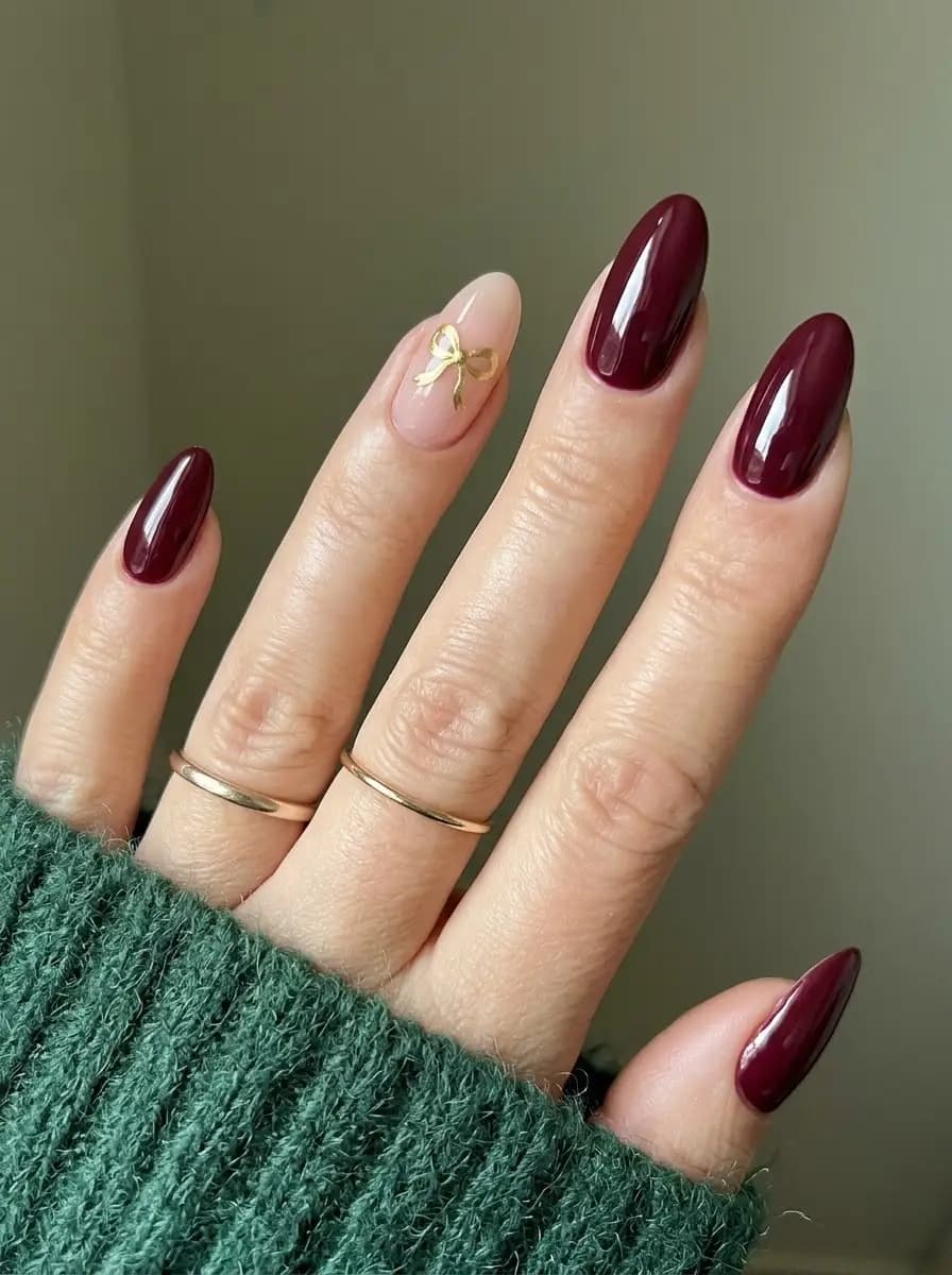 Deep burgundy Christmas nails with a delicate metallic-gold bow on a nude accent nail