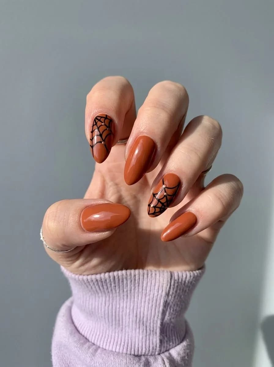 Short almond glossy pumpkin-orange nails with bold black spiderweb accents near the edges