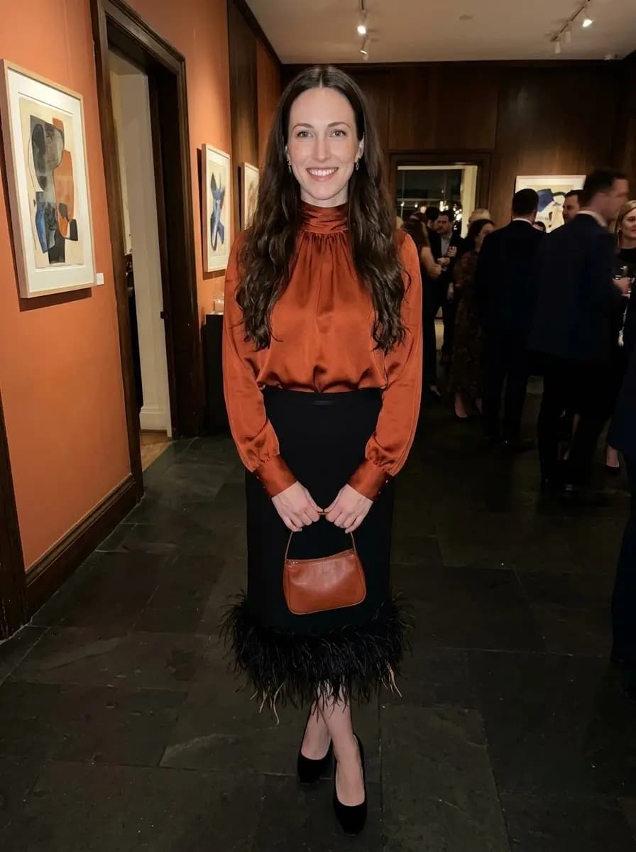 Gathered rust-colored satin high-neck blouse tucked into a black midi skirt with delicate black feather trimming